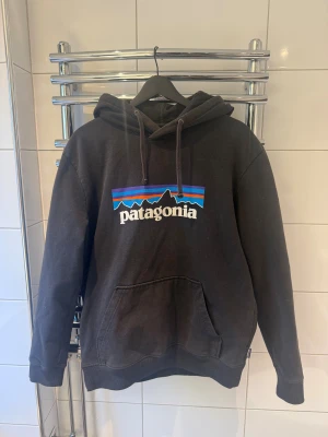 Svart Patagonia hoodie med tryck - Svart hoodie från Patagonia med klassiskt färgglatt logotryck på bröstet i bra skick. Köpt från Zalando, skriv för fler bilder. 