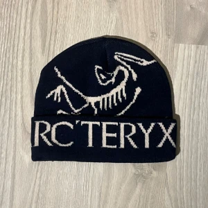 Mörkblå Arcteryx mössa - Snygg mörkblå mössa från Arcteryx med vit logga och stor text framtill. Stickad design som passar perfekt för kalla dagar. Stilren och sportig look som funkar till både street och outdoor.
