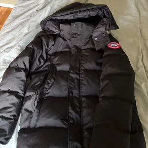 Svart pufferjacka från Canada Goose - Svart pufferjacka från Canada Goose med huva och klassisk logga på ärmen. Jackan har en quiltad design, dragkedja och knappar framtill. Perfekt för kalla dagar med sin tjocka och fluffiga look. Snygg och stilren modell som håller dig varm hela vintern.