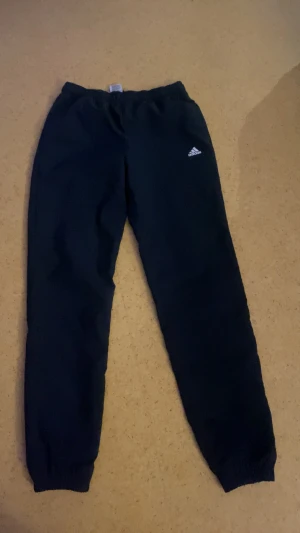 Svarta mjukisbyxor från Adidas - Svarta mjukisbyxor från Adidas med klassisk logga på vänster lår. Byxorna har elastisk midja och muddar vid bensluten för skön passform. Perfekta för chill eller träning, tillverkade i mjukt material som känns nice mot huden.