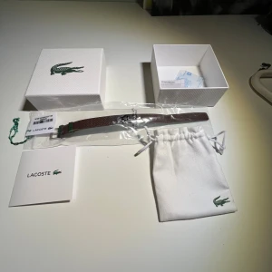 Brunt armband från Lacoste i skinn - Snyggt brunt armband från Lacoste i äkta skinn med texturerad yta och silverfärgad krokodillogga framtill. Armbandet har klassisk form och levereras med original ask och tygpåse med Lacoste-logga. Min handled är 17cm omkrets och armbandet passar perfekt på mig om man har den i sista hålet.Perfekt för dig som gillar stilrena accessoarer med premiumkänsla.