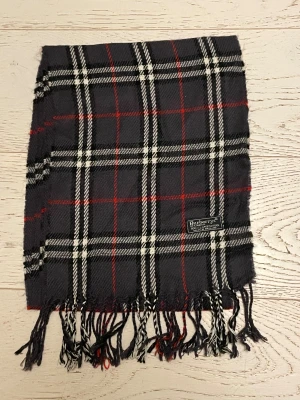 Rutig mörkgrå ullhalsduk Burberrys - Snygg halsduk från Burberrys i 100% lammull, tillverkad i England. Mörkgrå bas med klassiskt rutmönster i vitt och rött, samt fransar i ändarna. Perfekt för dig som vill ha en tidlös accessoar med lyxig känsla och ikonisk design.