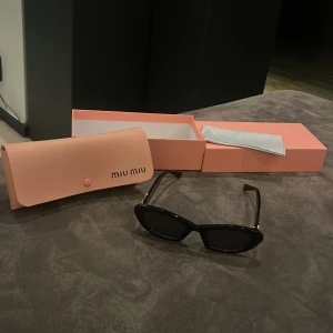 Svarta Miu Miu solglasögon med gulddetaljer - Snygga svarta solglasögon från Miu Miu med cool cat eye-form och exklusiva gulddetaljer vid skalmarna. Kommer med original rosa fodral och ask samt putsduk. Perfekt accessoar för att lyfta din stil och ge en lyxig vibe. ALLT INGÅR