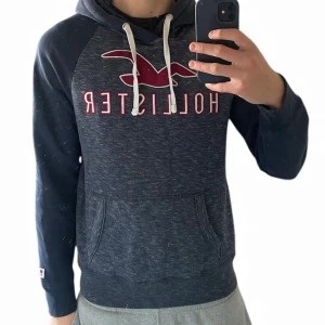Mörkblå hoodie från Hollister | Medium - Snygg mörkblå hoodie från Hollister med vit snörning och stort rött Hollister-tryck på bröstet. Tröjan har huva, långärmade raglanärmar och en praktisk magficka. Materialet är mjukt och skönt, perfekt för chill dagar. 