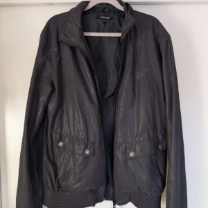 Svart bomberjacka från Junk de Luxe - Snygg svart bomberjacka från Junk de Luxe med glansig finish, dragkedja framtill och två stora fickor med knappar. Jackan har ribbade muddar, ståkrage och diskret broderad krona på bröstet. Perfekt för dig som gillar stilren och urban look.