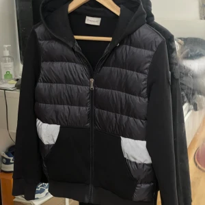 Svart pufferjacka med huva från Moncler - Svart pufferjacka från Moncler med huva och dragkedja framtill. Jackan har quiltade partier i glansigt material på framsidan och reflekterande detaljer vid fickorna. Ärmar och rygg är i mjuk bomull, och Moncler-logga sitter på vänster ärm. Perfekt för höst och vinter. Passar storlek S-XS 