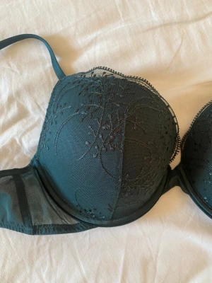 Grön bh från Primark - Supersnygg mörkgrön bh från Primark med broderade spetsdetaljer och lätt glansig yta. Axelbanden är justerbara och kuporna har ett elegant mönster. Push up bh med vaddering som är mjuk. Helt ny.  Storlek 80B europeisk storlek