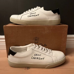 Saint Laurent vita sneakers i skinn - Snygga vita sneakers från Saint Laurent med handskriven logga på sidan. Skorna har rund tå, platt sula och svart detalj på hälen. Tillverkade i mjukt skinn med klassisk snörning och stilren design som passar till jeans eller dressat.