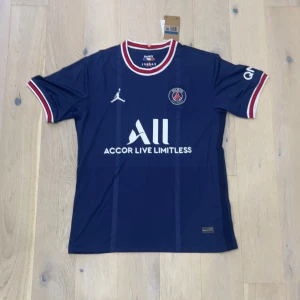 PSG marinblå fotbollströja Jordan - Snygg PSG fotbollströja i marinblått med vita och röda detaljer på krage och ärmslut. Tröjan har korta ärmar, PSG-logga på bröstet och Jordan-logga. Materialet är lätt och andas, perfekt för träning eller match. Tryck med 'Accor Live Limitless' på framsidan.