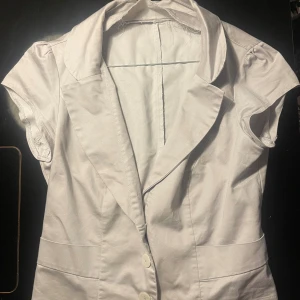 Vit kortärmad skjorta med knappar - Jättesöt ljusgrå blus med korta puffärmar och figursydd midja. Sitter jättefint men har en ganska stor urringning. Är i bra skick men lite sliten vid kragen, ingen som syns dock när man har på sig den.