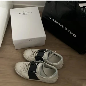 Valentino Garavani Open sneakers  - Säljer ett par Valentino Garavani Open sneakers i vitt skinn med bred svart rem över foten. Klassisk rund tå och platt sula med mönstrad undersida. Snygg och stilren design, perfekt för dig som gillar exklusiva sneakers med edge.