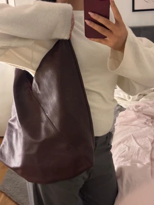 Stor vinröd axelväska - Säljer en stor och mjuk axelväska i vinrött skinn med enkel, stilren design. Väskan har ett brett handtag och en praktisk innerficka med dragkedja. Perfekt för dig som gillar minimalistisk look och vill ha plats för allt du behöver.