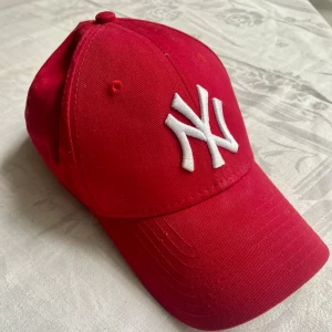 Röd New York Yankees keps - Snygg röd keps från New York Yankees med klassisk böjd skärm och broderad vit NY-logga framtill. Perfekt accessoar för att lyfta din streetwear-look. Justerbar passform baktill och tillverkad i bomull för skön känsla.