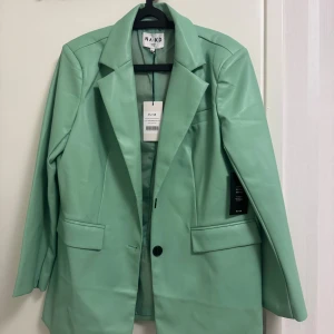 Mintgrön oversized kavaj från NA-KD - Maxi oversized kavaj i färgen dusty mint från NA-KD. Jackan har klassisk krage, långa ärmar, framfickor med lock och knäppning med en knapp. Materialet är syntetiskt PU som ger en cool skinnlook. Perfekt för dig som gillar statement-plagg och trendiga färger.