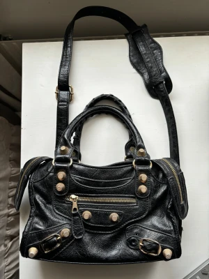 Vintage svart väska - Cool svart vintage  axelväska i mjukt skinn med guldiga detaljer och dragkedjor. Väskan har både handtag och justerbar axelrem, samt flera snygga nitar och spännen framtill. Perfekt storlek för det viktigaste och ger en edgy vibe till din outfit.