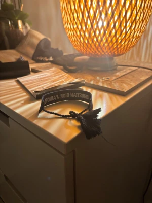 Christian Dior J'adior armband i svart - Snyggt svart armband från Christian Dior med texten 'J'adior' broderad i vitt. Armbandet har justerbar knytning med tofsar i ändarna och är gjort i ett mjukt tygmaterial. Perfekt accessoar för att lyfta din outfit med en lyxig touch. Helt nytt! Bara skriva vid frågor 