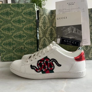 Gucci Ace sneakers med orm - Gucci Ace sneakers i vitt skinn med ikonisk broderad röd och svart orm på sidan, gröna och röda ränder samt metallicröd och grön häl med Gucci-logga. Klassisk rund tå, snörning och platt sula. Lyxig och unik detalj till din outfit. Dom är använda 1 gång. Kvitto och påsen följer med men om ni vill ha boxen då blir det 300 kr till. Men priser är inte hugget i sten kom med alla möjliga frågor
