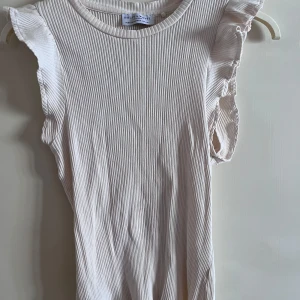 Ribbad ärmlös topp Holly & Whyte - Supersöt ribbad topp i ljusbeige från Holly & Whyte by Lindex. Modellen är ärmlös med volangdetaljer vid axlarna och rund halsringning. Perfekt för dig som gillar en feminin och stilren look. Materialet är mjukt och stretchigt, vilket gör den riktigt skön att bära.