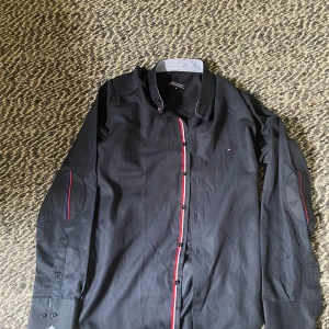 Svart skjorta från Tommy Hilfiger M - Svart skjorta från Tommy Hilfiger i bomull med lång ärm och coola detaljer som röda och blå ränder längs knappraden och på armbågarna. Klassisk krage med kontrast på insidan och små broderade loggor. Perfekt för dig som gillar stilrena plagg med en twist.