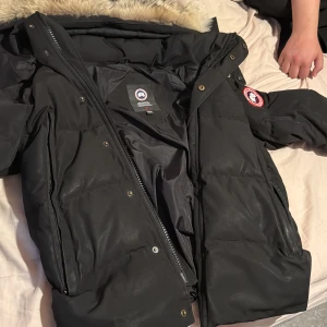 Svart dunjacka från Canada Goose - Svart dunjacka från Canada Goose med fluffig päls på huvan, klassisk logga på ärmen och dragkedja framtill. Jackan har lång ärm, flera fickor och är perfekt för kalla vinterdagar. Materialet är polyester och dun, med detaljer i päls på huvan.
