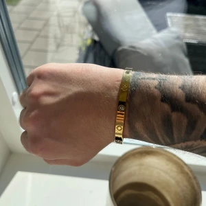 Guldigt armband med stenar - Snyggt och stilrent armband i guldton med blanka ytor och små infällda stenar. Armbandet har dekorativa detaljer med klöverformade mönster mellan stenarna och är smalt och elegant på handleden. Perfekt för dig som gillar att sticka ut med accessoarer. Oanvänd 
