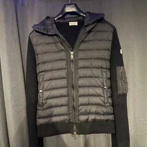 Moncler cardigan - Svart pufferjacka från Moncler med stickade ärmar och vadderad kropp. Jackan har huva, tvåvägsdragkedja och fickor med tryckknappar. Moncler-logga på ärmen och extra ficka med dragkedja på ena ärmen. Perfekt för kyliga dagar.
