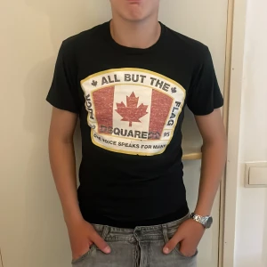 Svart Dsquared2 t-shirt med tryck - Säljer en svart t-shirt från Dsquared2 med stort tryck framtill som har ett rött lönnlöv och texten 'All But The Flag'. T-shirten är kortärmad och har en tight passform. Perfekt för dig som gillar streetwear och vill sticka ut med en snygg logga. Storlek S