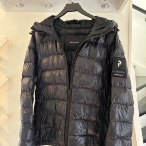 Svart dunjacka från Peak Performance  - Svart dunjacka från Peak Performance med glansig finish och klassisk quiltad design. Jackan har huva, dragkedja framtill och logotyp på ärmen. Perfekt för kalla dagar. Passar även M.
