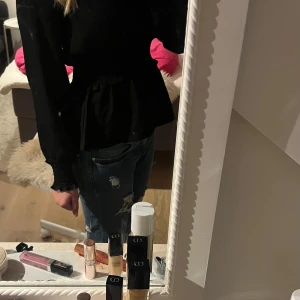 Svart blus med smock och volang - Säljer en svart blus med långärmade ärmar och smock i midjan och vid ärmsluten. Blusen har en volangkant nedtill och är tillverkad i ett mjukt, lätt material som ger en snygg siluett. Perfekt att styla med jeans eller kjol för en trendig look.