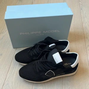 Philippe Model trainers - Svarta Philippe Model sneakers med vita detaljer och logga på sidan. Skorna har snörning, rund tå och platt sula. Ovandelen är i en mix av mocka och syntetmaterial, med vit hälkappa och svart/vit sula. Snygg och sportig design, perfekt för en trendig look. Använda 1 gång så 10/10 skick!