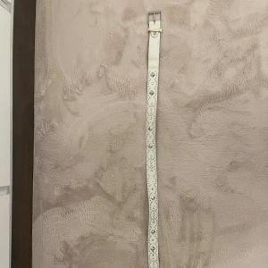 Vit bälte med nitar och hålmönster - Säljer ett vitt bälte med coola silverfärgade nitar och dekorativa hål längs hela remmen. Bältet har en klassisk metallspänne och är gjort i skinnimitation. Perfekt för att ge din outfit lite edge och detaljer.