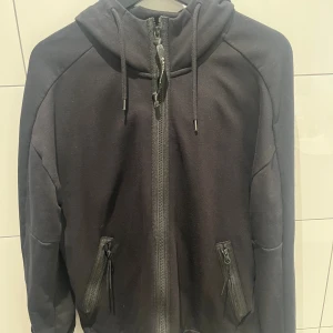 Svart hoodiejacka från C.P. Company - Svart hoodiejacka från C.P. Company med ikoniska goggles i huvan. Jackan har dragkedja framtill, två fickor med dragkedja och justerbar huva med snörning. Tillverkad i mjuk bomull för en skön och avslappnad look. Perfekt för dig som vill sticka ut med streetwear-vibe. Köpt för 3700 säljer för 900 kr.