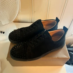 Christian Louboutin svarta sneakers med nitar - Svarta sneakers från Christian Louboutin i mocka med coola svarta nitar på tån och klassisk röd sula. Skorna har snörning och rund tå, och kommer med originalkartong och dustbag. Perfekta för dig som vill sticka ut med en lyxig stil. Lite förstörda i sulan vid hälen och kådan har ett litet hål