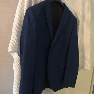 Blå kavaj från Jack & Jones - Snygg marinblå kavaj från Jack & Jones Premium med klassisk passform. Kavajen har två knappar framtill, fickor med lock och dekorativa knappar vid ärmslutet. Perfekt för dig som vill ha en stilren och modern look. Nypris var 600, använd endast 1 gång