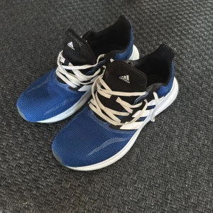 Blåa Adidas sneakers med vit sula - Snygga blåa Adidas sneakers med vit sula och svarta detaljer. Skorna har vit snörning och är tillverkade i ett ventilerande syntetmaterial. Perfekta för dig som gillar sportig stil och vill ha bekväma skor till vardagen.