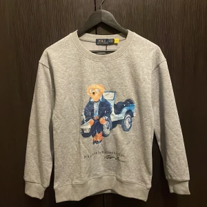 Grå sweatshirt Polo Ralph Lauren S - Grå sweatshirt från Polo Ralph Lauren med ikoniskt björntryck framtill. Tröjan har rund hals, ribbade muddar och är tillverkad i mjuk bomullsmix. Perfekt för dig som gillar streetstyle och vill ha något med coolt print.
