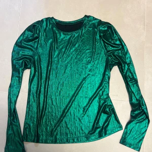 Grön glittrig långärmad topp - Säljer en grön glittrig långärmad topp med rund halsringning. Tyget har en metallic finish som verkligen fångar ljuset och ger en cool effekt. Perfekt för dig som vill sticka ut med en färgstark och trendig look.