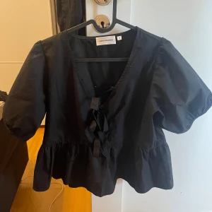 Svart blus med knyt från Carin Wester - Svart blus från Carin Wester med v-ringning och dekorativ knytning framtill. Modellen har korta puffärmar och volang nertill som ger en trendig siluett. Perfekt för dig som gillar stilrena och coola plagg med det lilla extra.