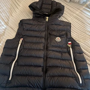 Svart dunväst från Moncler med huva - Säljer en mörkblå dunväst från Moncler med huva och vita dragkedjor. Västen har en bröstficka med Moncler-logga, två snedställda fickor med dragkedja och quiltad design. Pris kan diskuteras och är öppen för byten.
