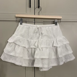 Vit volangkjol från Gina Tricot XS - Supersöt vit kortkjol från Gina Tricot i storlek XS. Kjolen har broderade detaljer, flera lager volanger och en dragsko i midjan. Perfekt för dig som gillar romantisk och feminin stil. Tillverkad i bomull för en luftig känsla. Aldrig använd, ordinarie pris 300kr