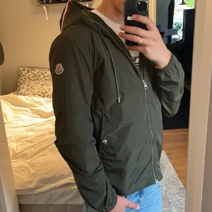 Moncler Grimperus KVITTO FINNS! - Säljer en snygg grön windbreaker från Moncler med huva och dragkedja framtill. Jackan har två fickor med knappar och Moncler logga på armen. Kvitto kommer med. Priset är inte hugget i sten.