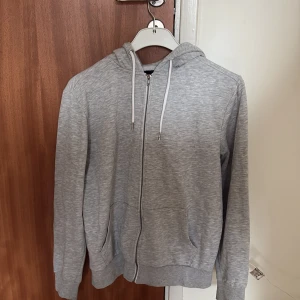 Grå hoodie med dragkedja och huva - En stilren grå hoodie med dragkedja framtill och vita snören i huvan. Tröjan har långa ärmar, ribbade muddar och två fickor på sidorna. Perfekt för en avslappnad look och enkel att matcha med andra plagg.