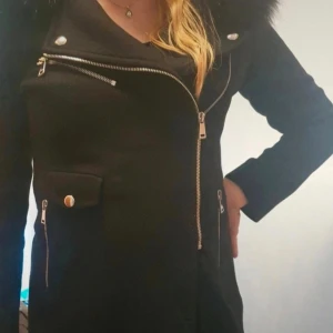 Svart kappa med fuskpäls från Zara - Svart kappa från Zara med stor krage i fuskpäls, snedställda dragkedjor och dekorativa knappar. Jackan har lång ärm, markerad midja och flera fickor framtill. Materialet är 75% ull och 25% polyamid, vilket gör den både varm och stilren.