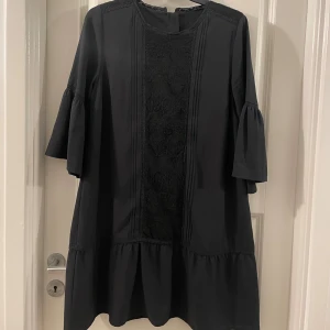 Svart kortklänning från Åhléns med spets - Svart kortklänning från Åhléns med trekvartsärmar och volangdetalj nedtill. Framsidan har dekorativ spets och diskreta veck, baksidan har en rad klädda knappar. Perfekt för dig som gillar en stilren look med fina detaljer.