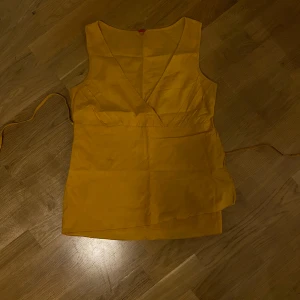 Gul omlottopp med knytband - Snygg gul ärmlös omlottopp med djup v-ringning och knytband i midjan. Toppen har en enkel och stilren design med lätt figursydd passform. Perfekt för att addera färg till din outfit.