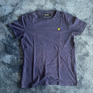 Marinblå t-shirt från Lyle & Scott - Snygg marinblå t-shirt från Lyle & Scott med klassisk rund hals och korta ärmar. På bröstet sitter den ikoniska gula örnloggan som ger en stilren touch. Tillverkad i mjuk bomull för skön komfort hela dagen.