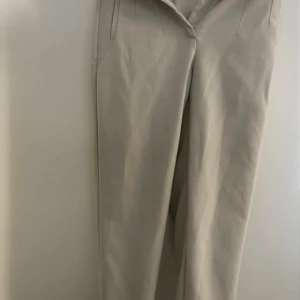 Beiga kostymbyxor från Zara - Stilrena beiga kostymbyxor från Zara med hög midja och raka ben. Byxorna har diskreta fickor och stängs med dragkedja, hake och knapp. Perfekta för en clean och snygg look. Materialet är mjukt och känns bekvämt mot huden.