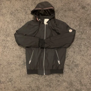 Svart Moncler vindjacka med huva - Storlek S sitter jäkligt bra 👍 den är i nyskick. Finns i Stockholm annars frakt                                 Svart vindjacka från Moncler med huva och snörning, hel dragkedja framtill och två sidofickor med dragkedja. Jackan har ribbade muddar och ett broderat Moncler-märke på ärmen. Insidan är fodrad med glansigt material och har en cool etikett med serietidningsmotiv.