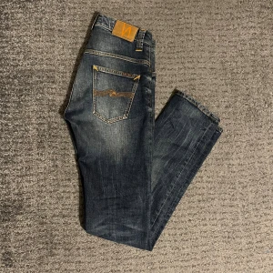 Blåa nudie jeans - Mörkblå jeans från Nudie Jeans med snygga slitningar och tydliga orange kontrastsömmar. Den har dessutom logga på bakfickan och läderpatch i midjan. Jeansen har rak passform. Storlek 30/34 