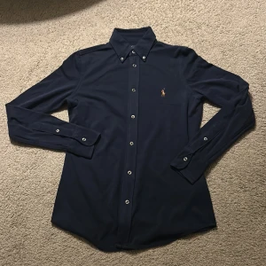 Mörkblå skjorta från Ralph Lauren - Snygg mörkblå skjorta från Ralph Lauren med klassisk krage och broderad logga på bröstet. Skjortan är i bomull, har långa ärmar och knäppning framtill. Perfekt för dig som gillar stilrena och tidlösa plagg.